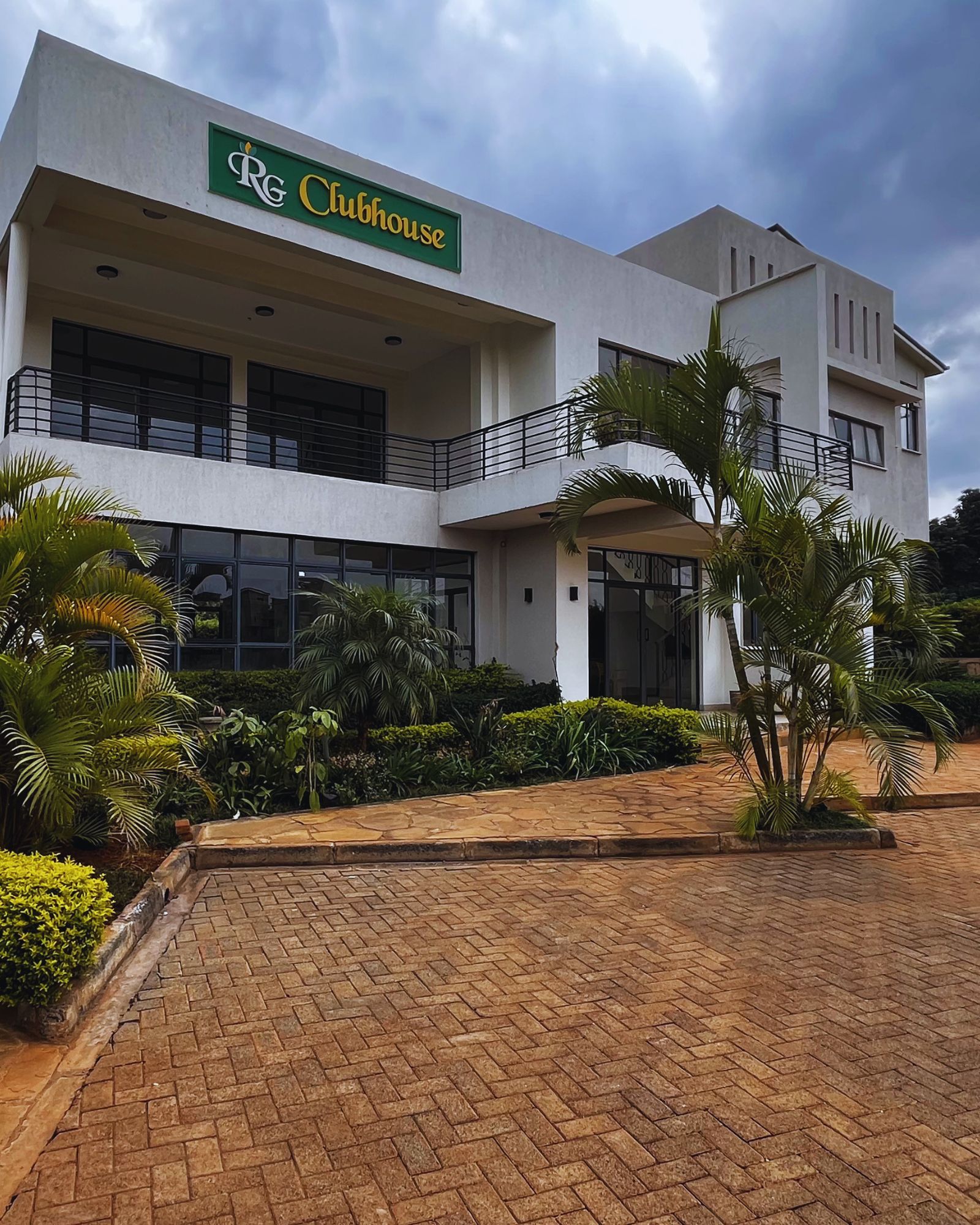 Two story RG Clubhouse entrance with balcony and green RG Clubhouse sign, flanked by palm trees and manicured hedges, paved driveway in front, overcast sky above, calm welcoming residential atmosphere